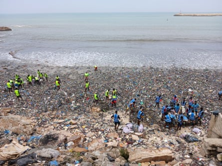 Wide shot shows a strip of beach covered in waste clothing with a huge team of workers in hi-vis dwarfed by the scale of the problem.