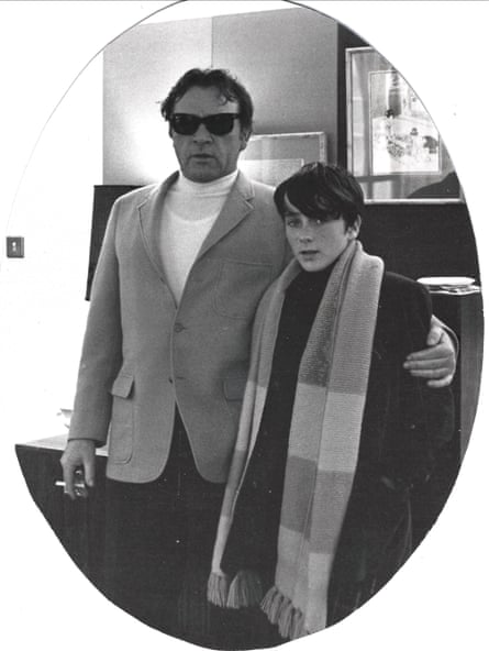 Richard Burton, wearing a whit polo neck top and a sports jacket, stands cigarette in hand, with his arm around Christopher who is wearing a black coat and a long stripy scarf, c 1969.