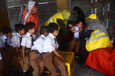 Young boys wearing brown trousers, white shirts and striped ties sit on a bench and watch while female health workers give a little boy an injection.