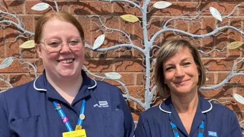 Adele Pavier and Ashleigh Robinson wearing dark blue NHS nurses uniforms, with lighter blue lanyards around their necks. They are smiling into the camera and standing in front of a metallic sculpture of a tree with different coloured leaves, each bearing an inscription.