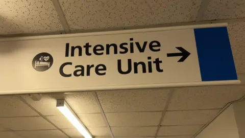 A white sign suspended from a hospital corridor ceiling. "Intensive Care Unit" is written in large black letters, with a black arrow pointing to the right.