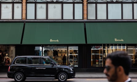 A London cab passes by two display case windows with awnings that read “Harrods” 