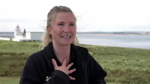 Sophia Jackson, who has blonde hair, holds her hand to her chest looking happy. A white lighthouse is in background. 