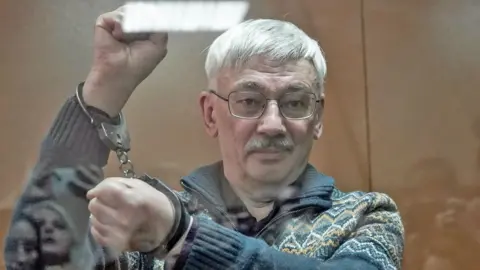 Reuters Oleg Orlov pictured behind the glass of a court dock, holding his arms in the air to show he is wearing handcuffs and clenching his first with a slight smile on his face. He wears a thick patterned jumper with a zip and collar, he has silver hair, a moustache and wire-rimmed glasses.