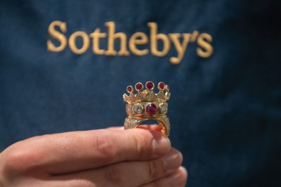 An unmanicured hand holds a ring that has a crown on top. It is gold and encrusted with diamonds and rubies. The Sotheby's logo is visible in the background. 