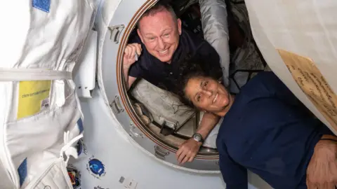 NASA Butch and Suni on space station - they are both smiling down at the camera in a circular capsule and appear to be in a gravity suspended environment. They are both dressed casually and the walls of the capsule appear to be made from a fabric-type material.  