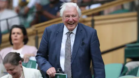 Getty Images Sir David Attenborough at Wimbledon. He is smiling as he takes his seat, and is wearing a dark blue-grey jacket and and black and white patterned tie