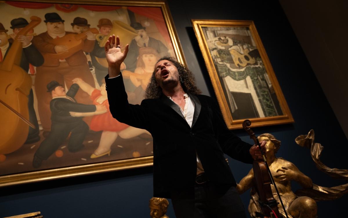 A man holding a violin is singing with his eyes closed and one hand raised, standing in front of two beautiful paintings of instruments