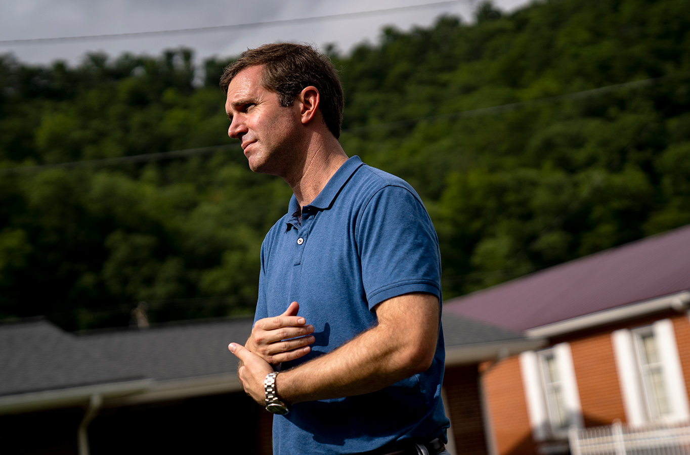 A man in profile looking in front of him and clasping his arms. The man is Kentucky Governor Andy Beshear and he is speaking to the press.