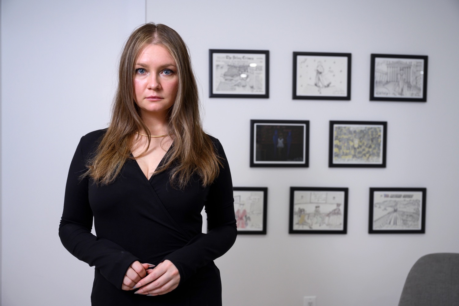 A woman wearing a black dress looks at the camera in front of eight framed drawings.