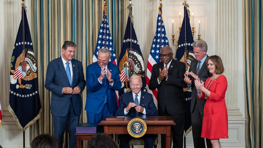 President Biden sits at a desk surrounded by other politicians and signs a bill