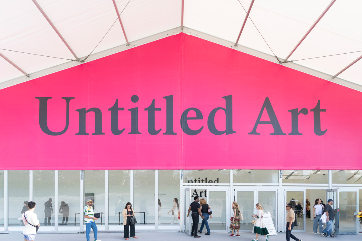 View of a tent with a pink banner that reads in black text 'Untitled Art'; people walk about below.