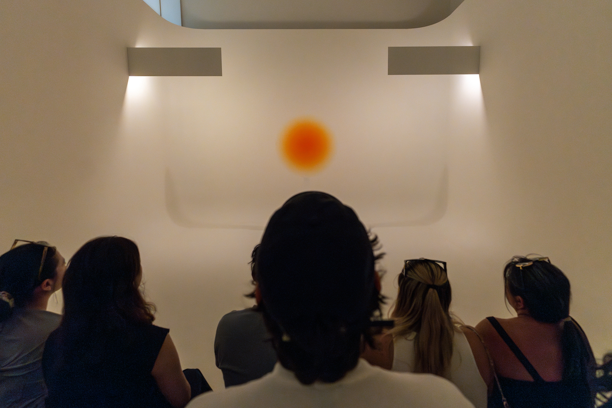 A crowd looks on at a yellow-org circle in a white cylindrical background