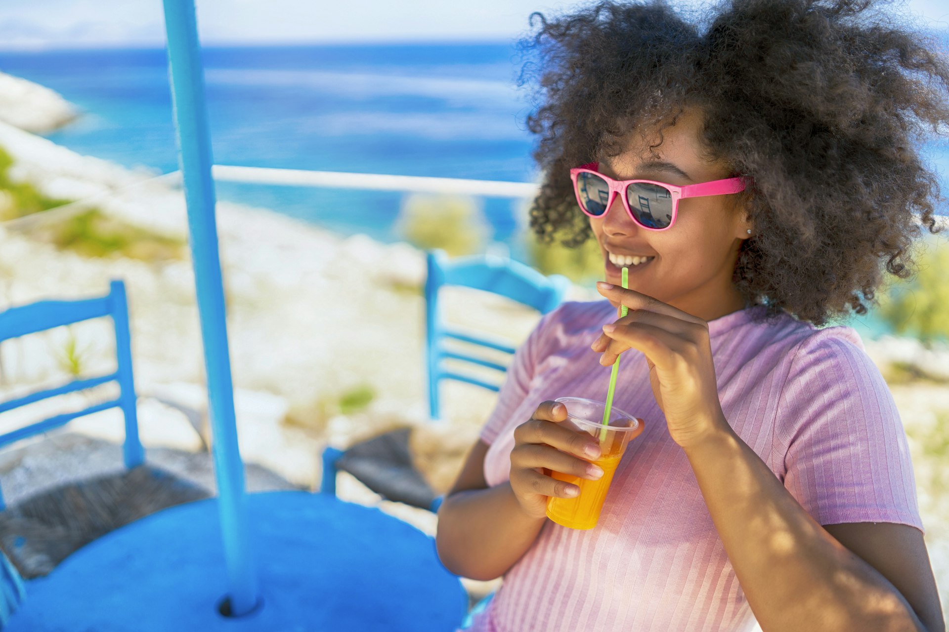 A woman is sitting in the sunshine smiling and drinking from a glass