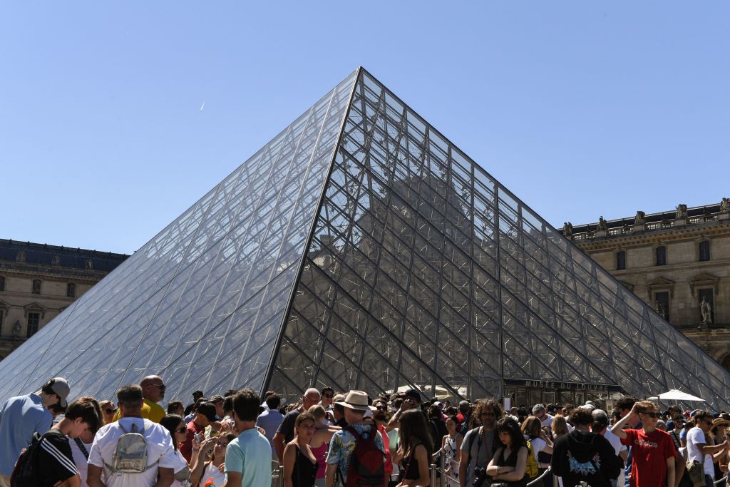 A crowd of people outside next to a large glass pyramid structure against a blue sky.