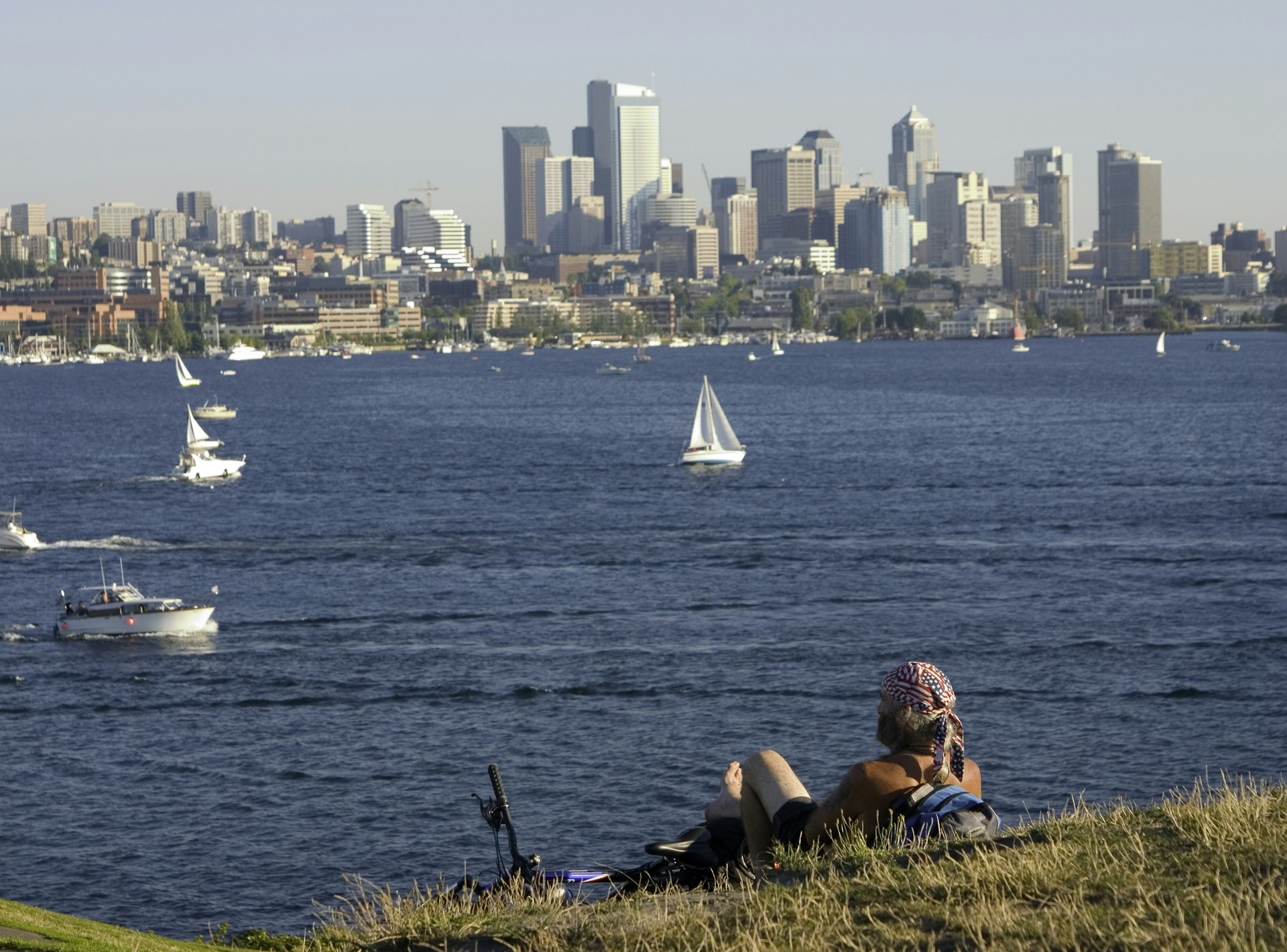 A person lies on grass looking out over a lake with boats on it. The city skyline is on the other side of the lake