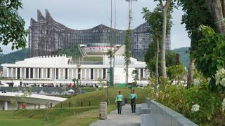 Two men walk along a pathway with large construction visible in the background