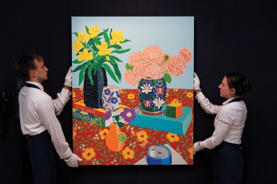 Two art handlers wearing gloves hold a painting that is a still life of various vases, some with flowers. There are two books (on Stuart Davis and one van Gogh) on the table, which has a floral table cloth. 