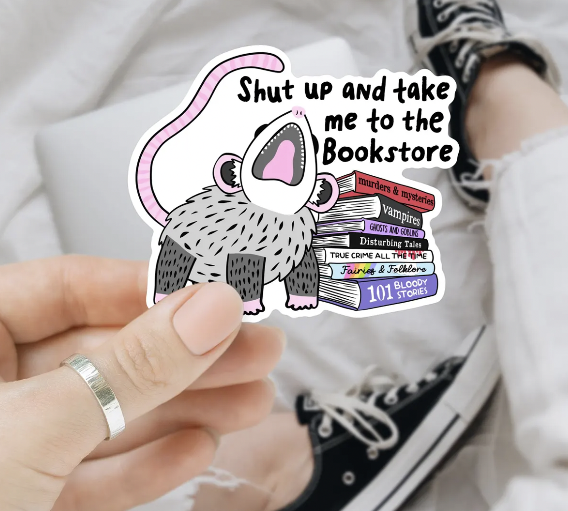 Image of a hand holding a vinyl sticker with an illustration of an opossum next to a stack of books saying "shut up and take me to the bookstore"