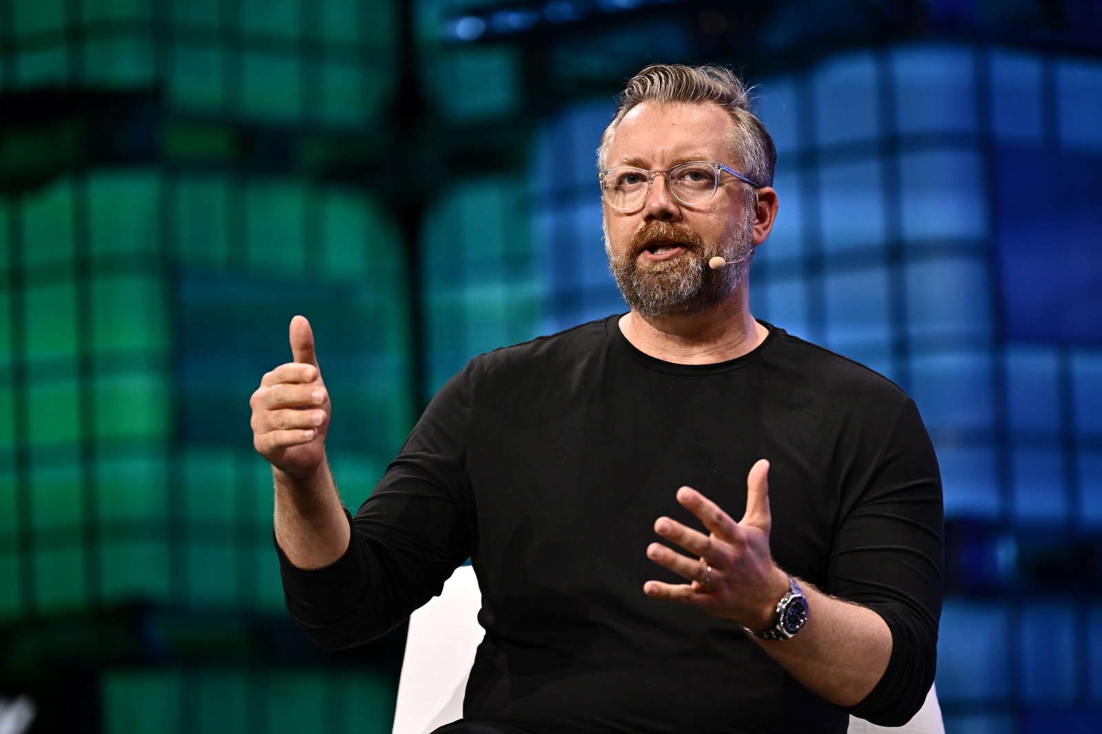 A man with short hair and glasses, in a long-sleeve black shirt, speaks into a headset at a conference