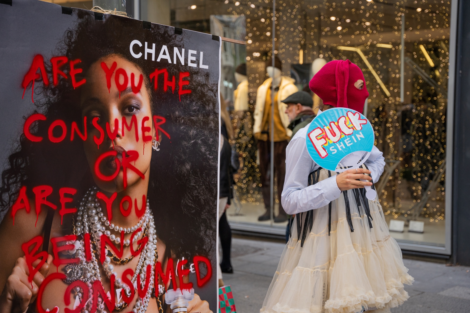 A person in a tutu and red balaclava holds a Shein-branded fan that has been painted over to say "FUCK Shein". An advertisement in the foreground warns passer-bys about consumption.