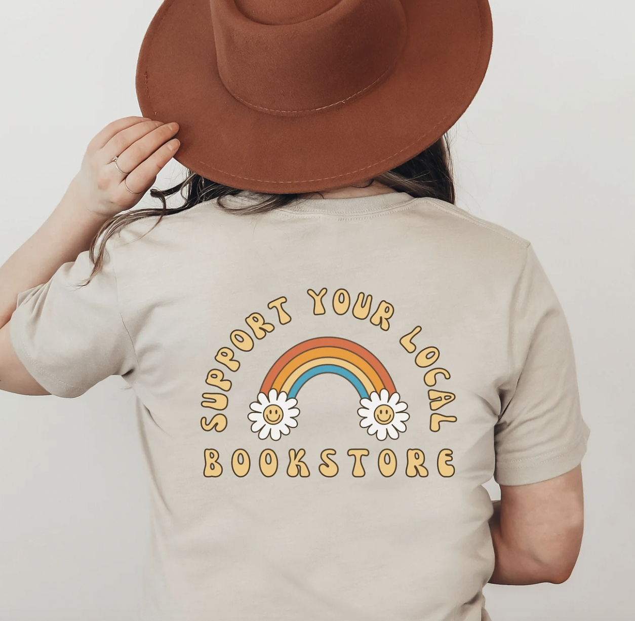 Image of someone's back wearing a taupe t-shirt with a rainbow and the phrase "support your local bookstore" on the back of the shirt