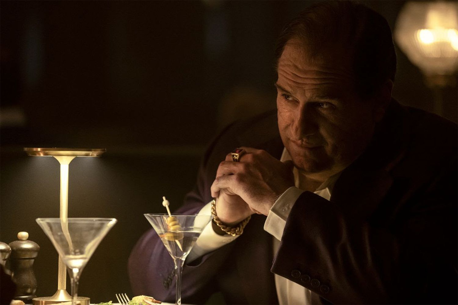 A man with a visible facial scar in a purple suit folding his hands in front of table with cocktail glasses in The Penguin.