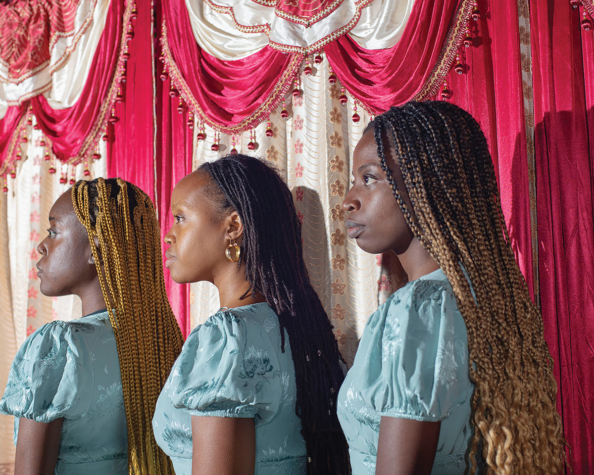 A horizontal photograph shows three women with brown skin and hair standing behind each other and looking off to the left. Behind them is a pink and white curtain.