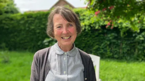 Lesley Clarke in her garden on a sunny day. She is smiling into the camera and wearing a blouse with thin vertical stripes and a grey cardigan. The grass and high hedge in the background are blurred.