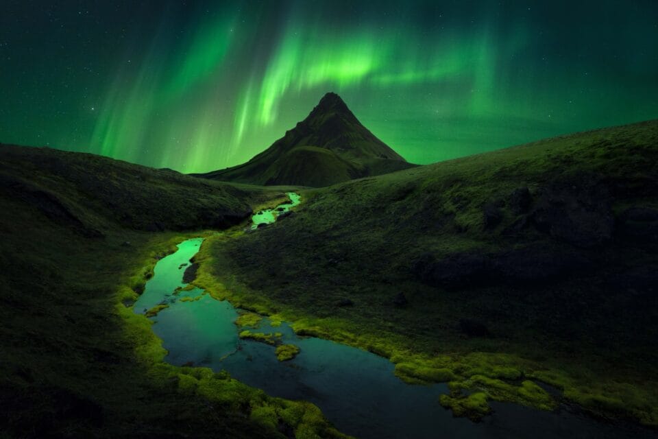 bright green lights shoot above a mountain with a small stream running to the foreground