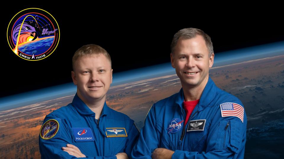  Two men in blue flight suits pose for a portrait with the arms crossed in front of a background of earth as seen from space. 