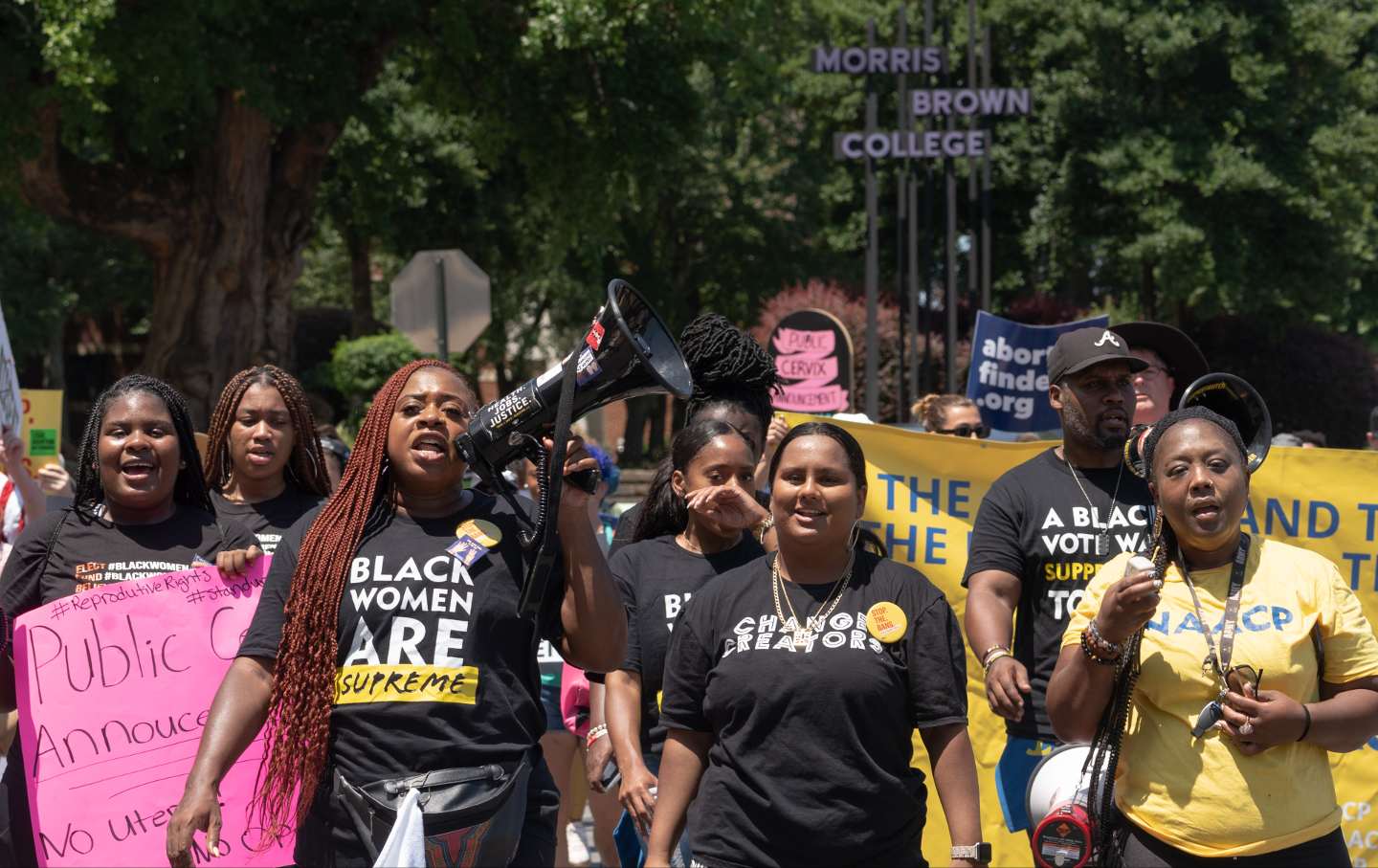 A woman wearing a t-shirt reading "Black Women Are Supreme" and holding a bullhorn marches with a crowd of protesters.