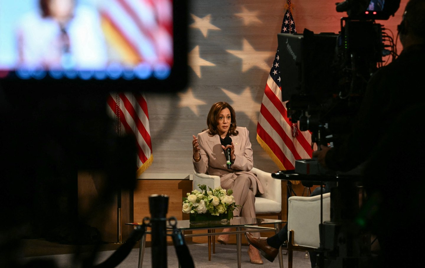 A wide-angle view of Kamala Harris sitting on a stage with a microphone, with a camera and screen in the foreground.