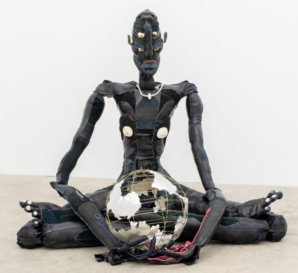 a lanky figure in black fabric sits on the floor cradling a globe-like sculpture