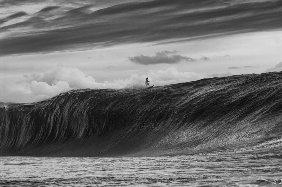 in a black and white photo, a large barreling wave begins to crash down on itself as a surfer rides atop it