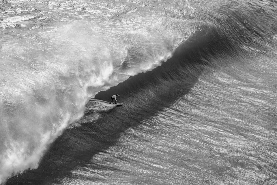 in a black and white photo, a large barreling wave begins to crash down on itself as a surfer rides the channel underneath