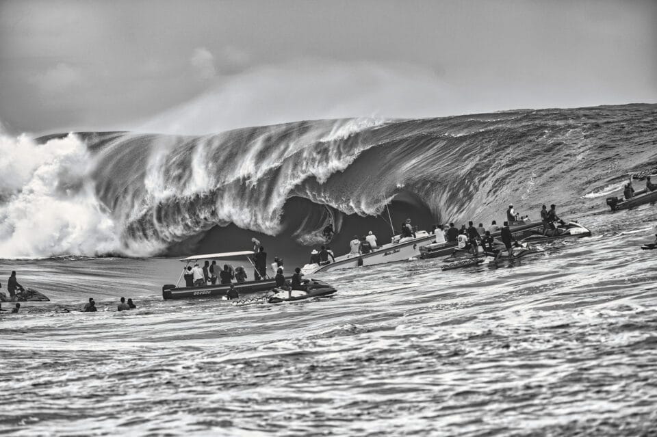 in a black and white photo, a large barreling wave begins to crash down on itself as groups of individuals in the water nearby watch