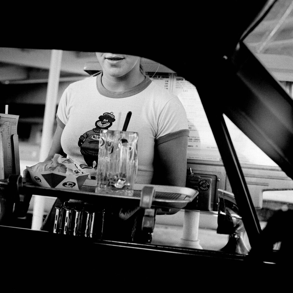 a black and white photograph of a waitress approaching a car serving a tray of food at a drive-in