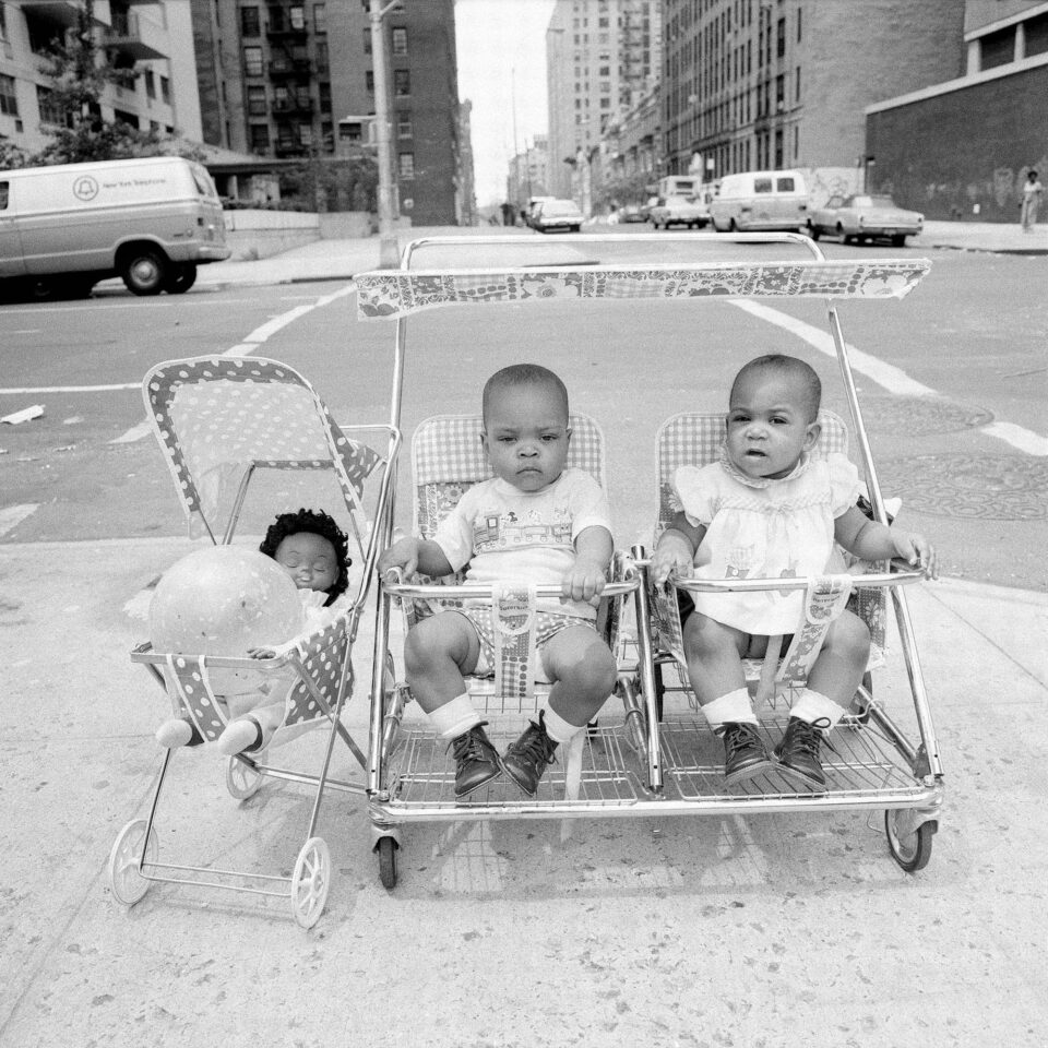 a black and white photograph of twins in a stroller next to another stroller with a doll in it