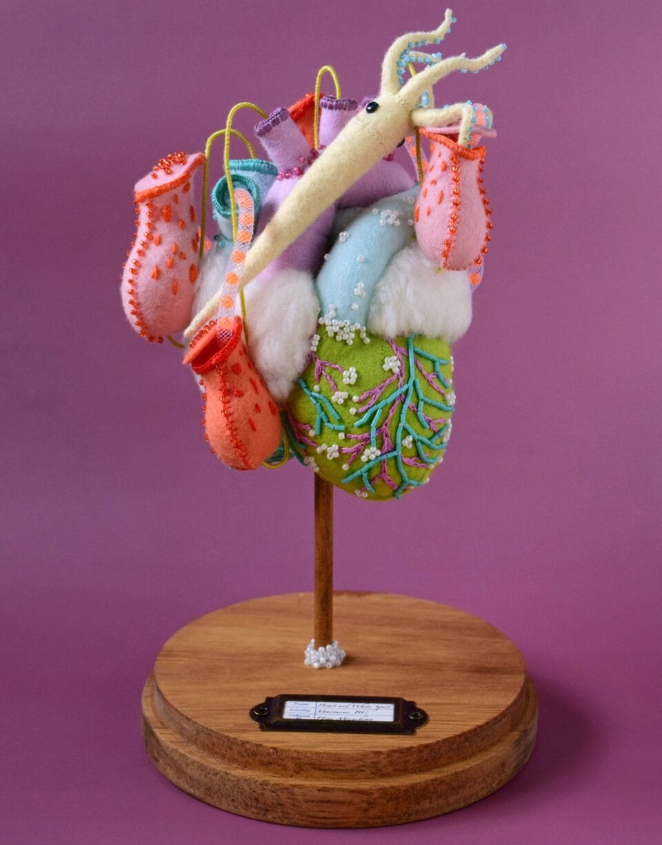 a white squid sprawls across a beaded green heart, which  stands on a wooden pedestal. there are small pink and red beaded arteries dangling from the organ