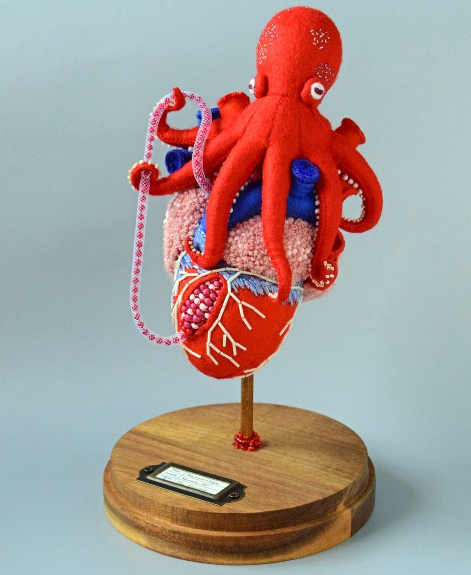 a red octopus sprawls across a beaded red and pink heart, which  stands on a wooden pedestal