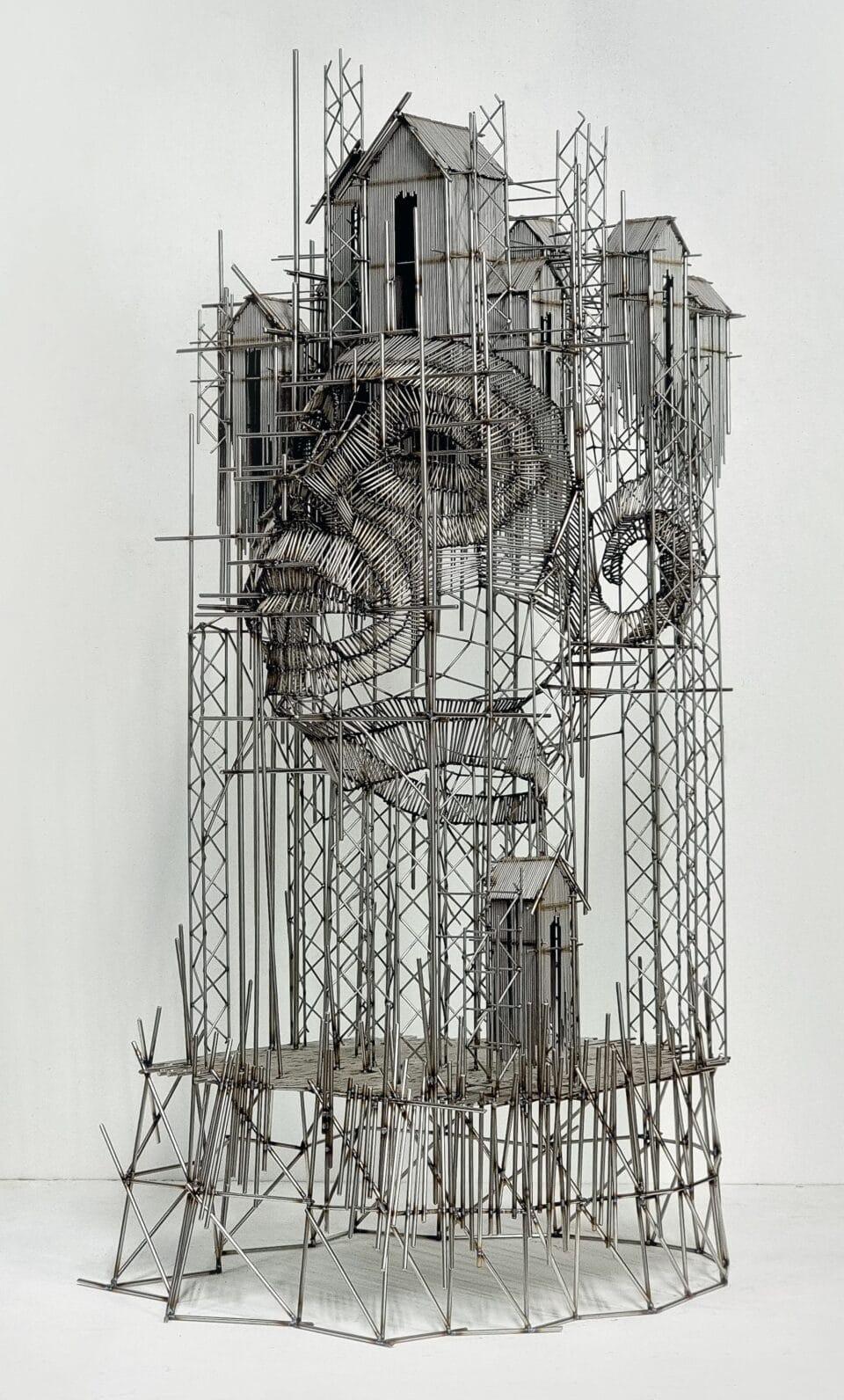 a wall sculpture made of numerous pieces of steel wire assembled in an abstract composition of house-like forms on scaffolding and the outline of a face