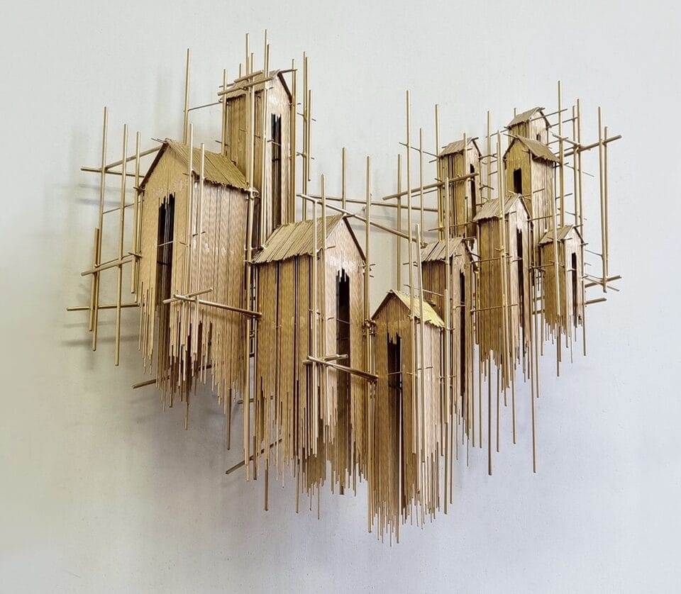 a wall sculpture made of numerous pieces of steel wire assembled in an abstract composition of house-like forms on gold scaffolding