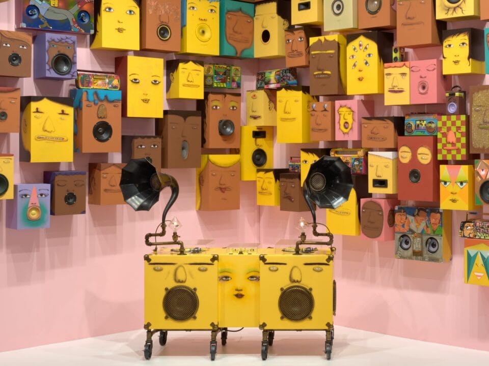 a detail of a collection of speakers painted with yellow and brown faces against a pink wall. a gramophone painted similarly stands in the front