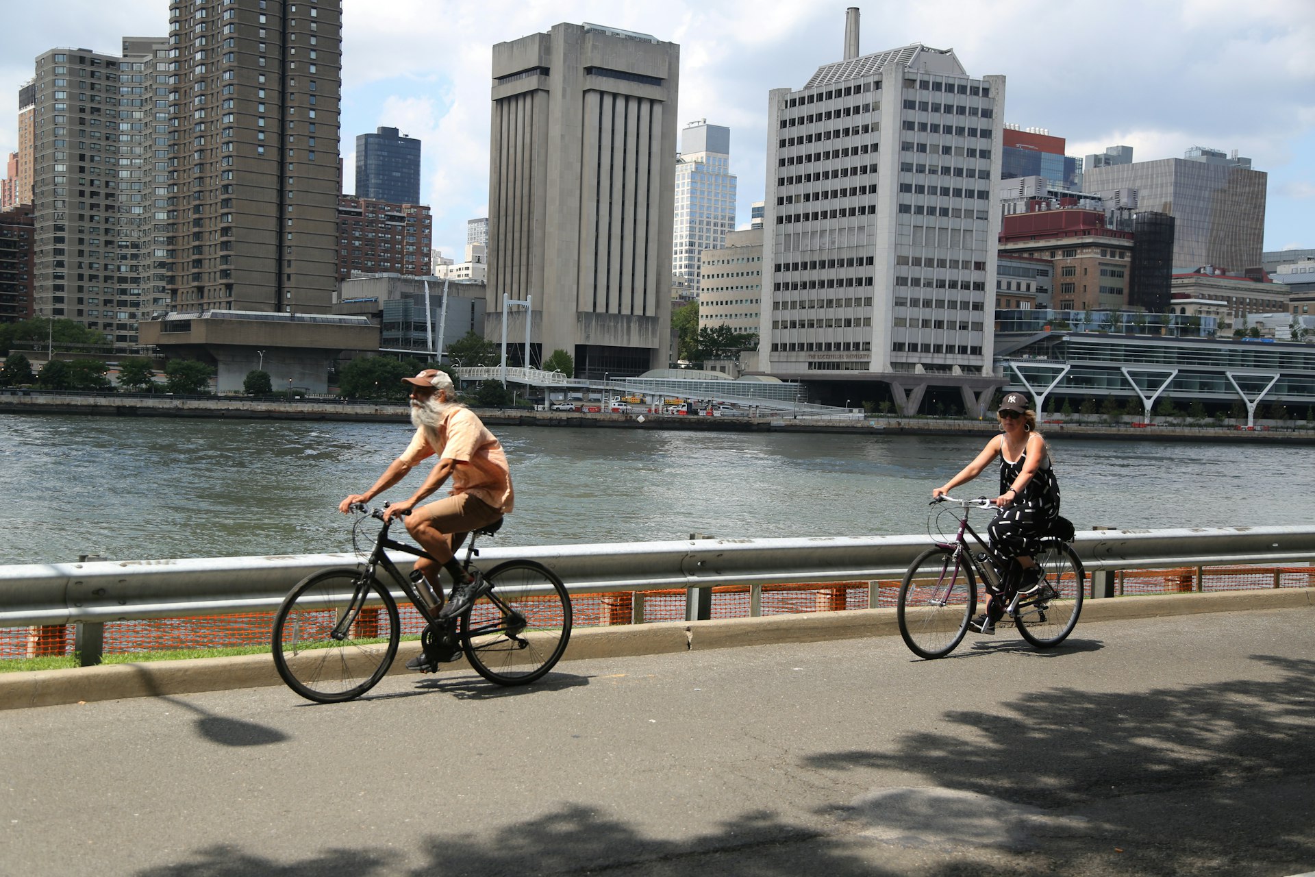 Two cyclists pedal along a path on the west side of Roosevelt Island, looking across the East River to the towers of Manhattan, New York City