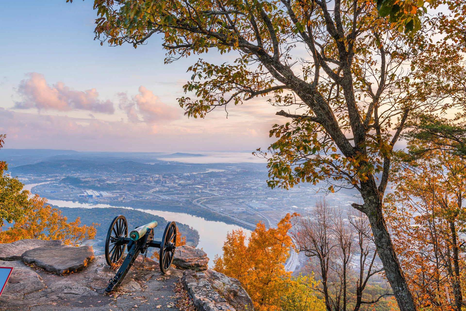 An old-style canon sits on a cliff edge pointing towards a city on the curve of a river. All the leaves on the trees in the surrounding state park are in reds and golds of autumn