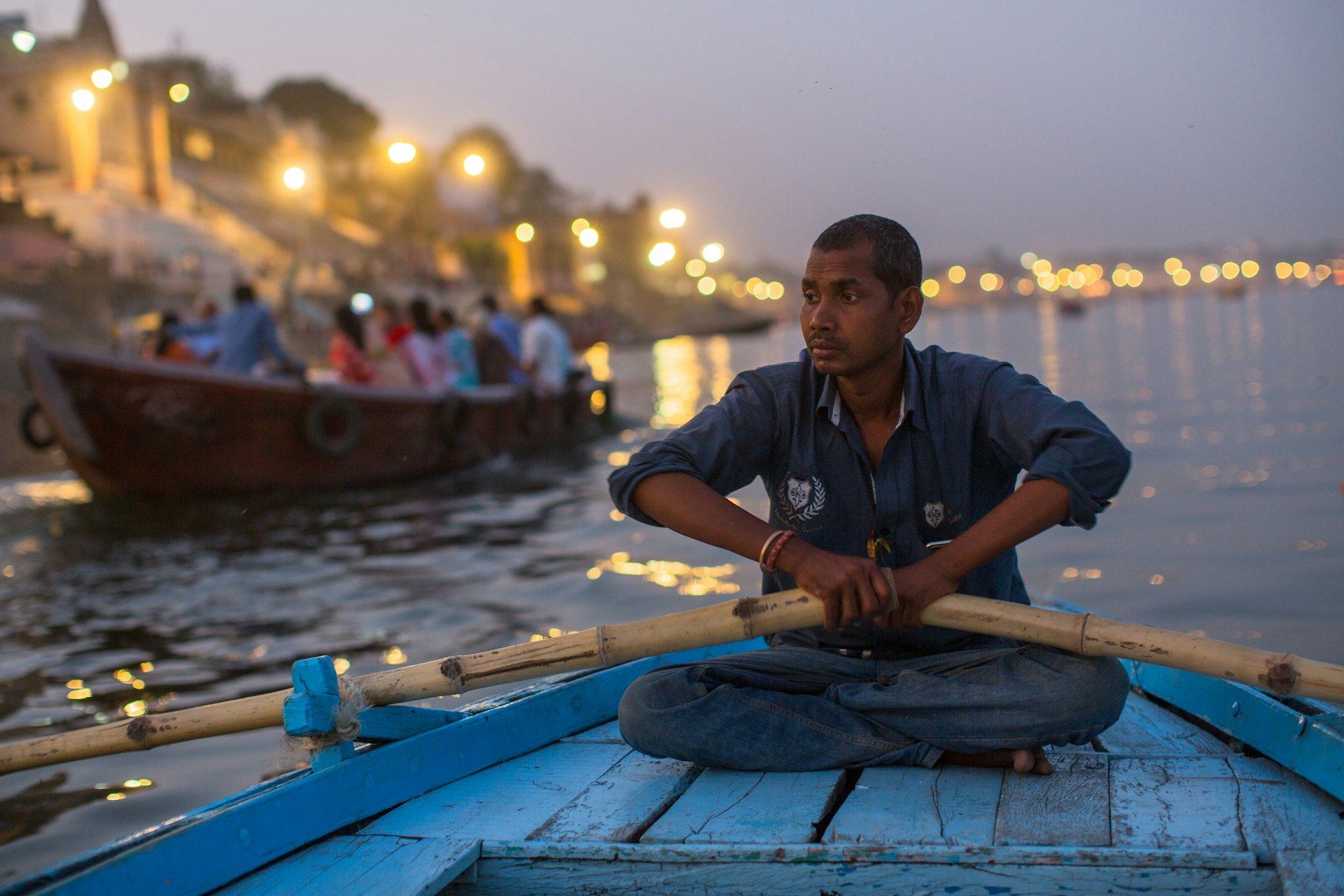 Close-up of a boatmen holding oars on the Ganga River at night with strings of lights in the background, Varanasi, India