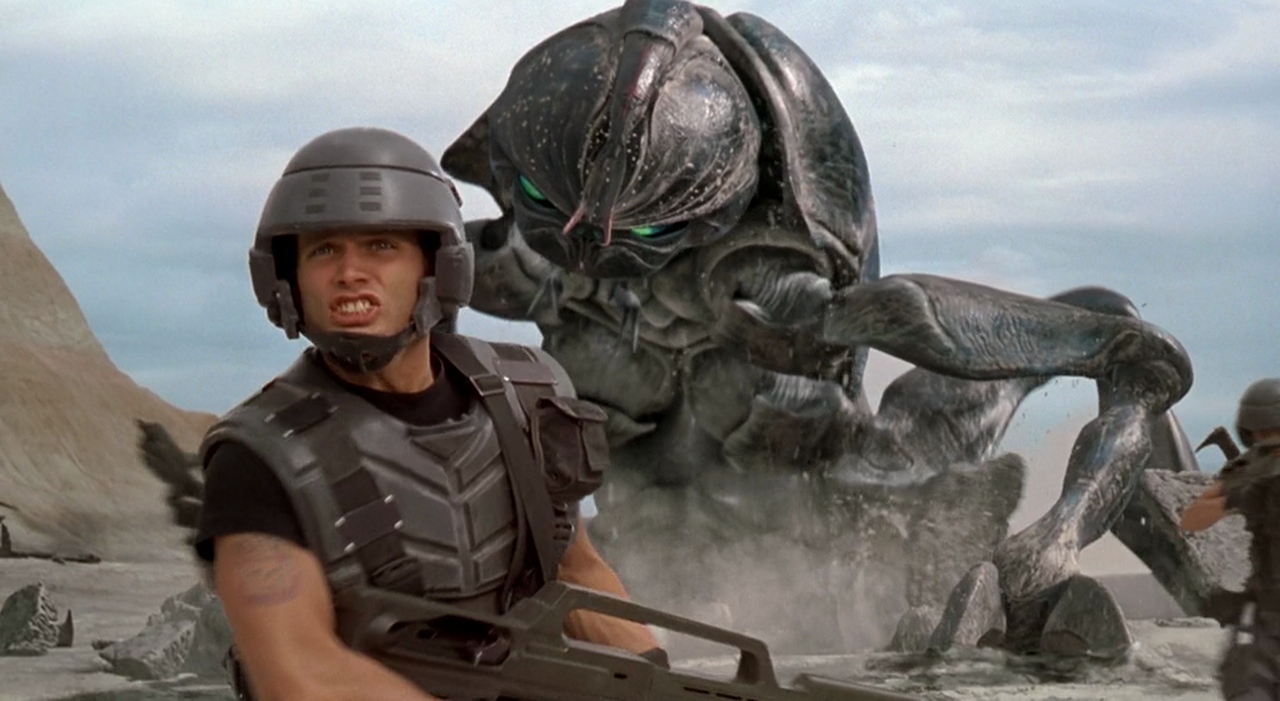 A man in soldier armor standing in front of a gigantic insect in Starship Troopers.